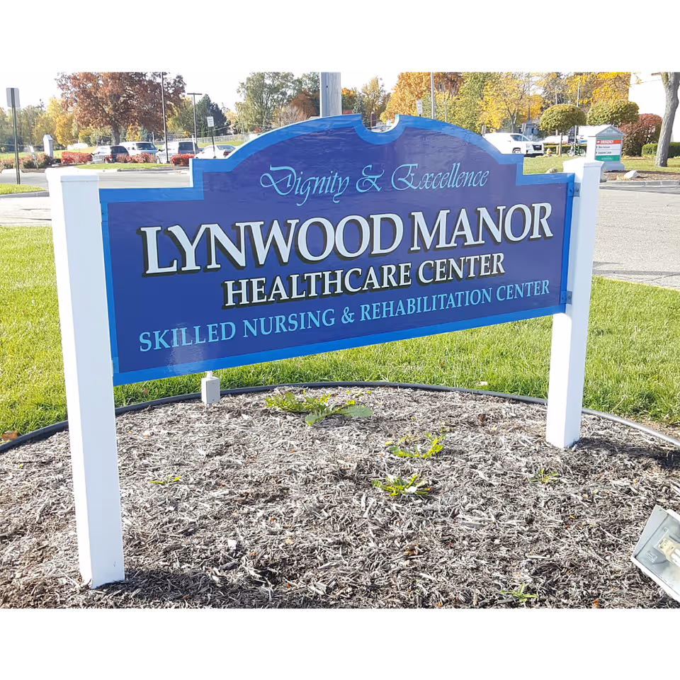 A blue sign with white posts in a landscaped area with mulch and small plants. The sign reads 'Dignity & Excellence LYNWOOD MANOR HEALTHCARE CENTER SKILLED NURSING & REHABILITATION CENTER'. In the background, there is a parking lot with cars and trees with autumn foliage.
