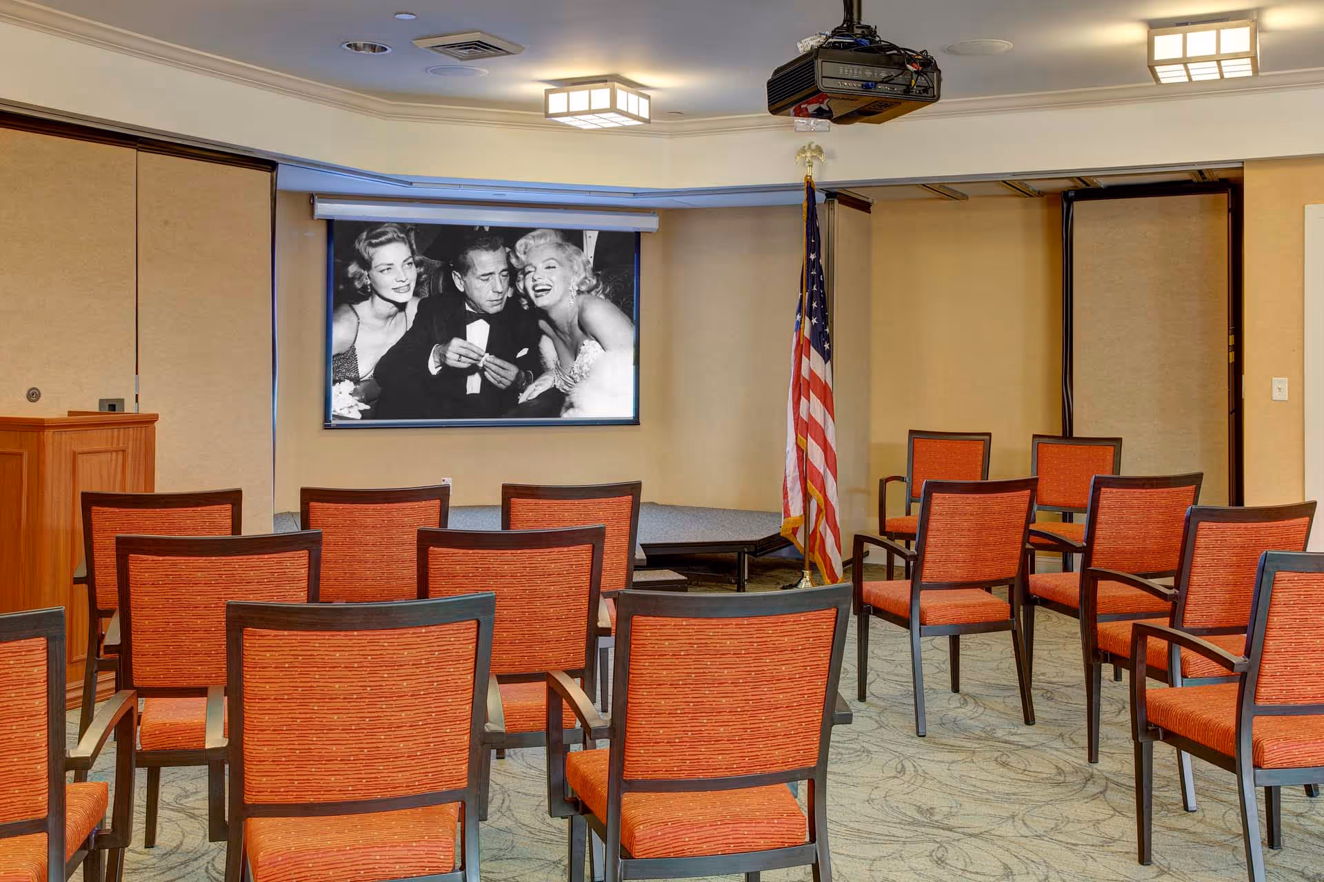 A small conference or presentation room with rows of orange cushioned chairs facing a small stage. On the stage, there is a black and white photo of three people displayed on a screen. An American flag stands to the right of the stage, and a projector is mounted on the ceiling.
