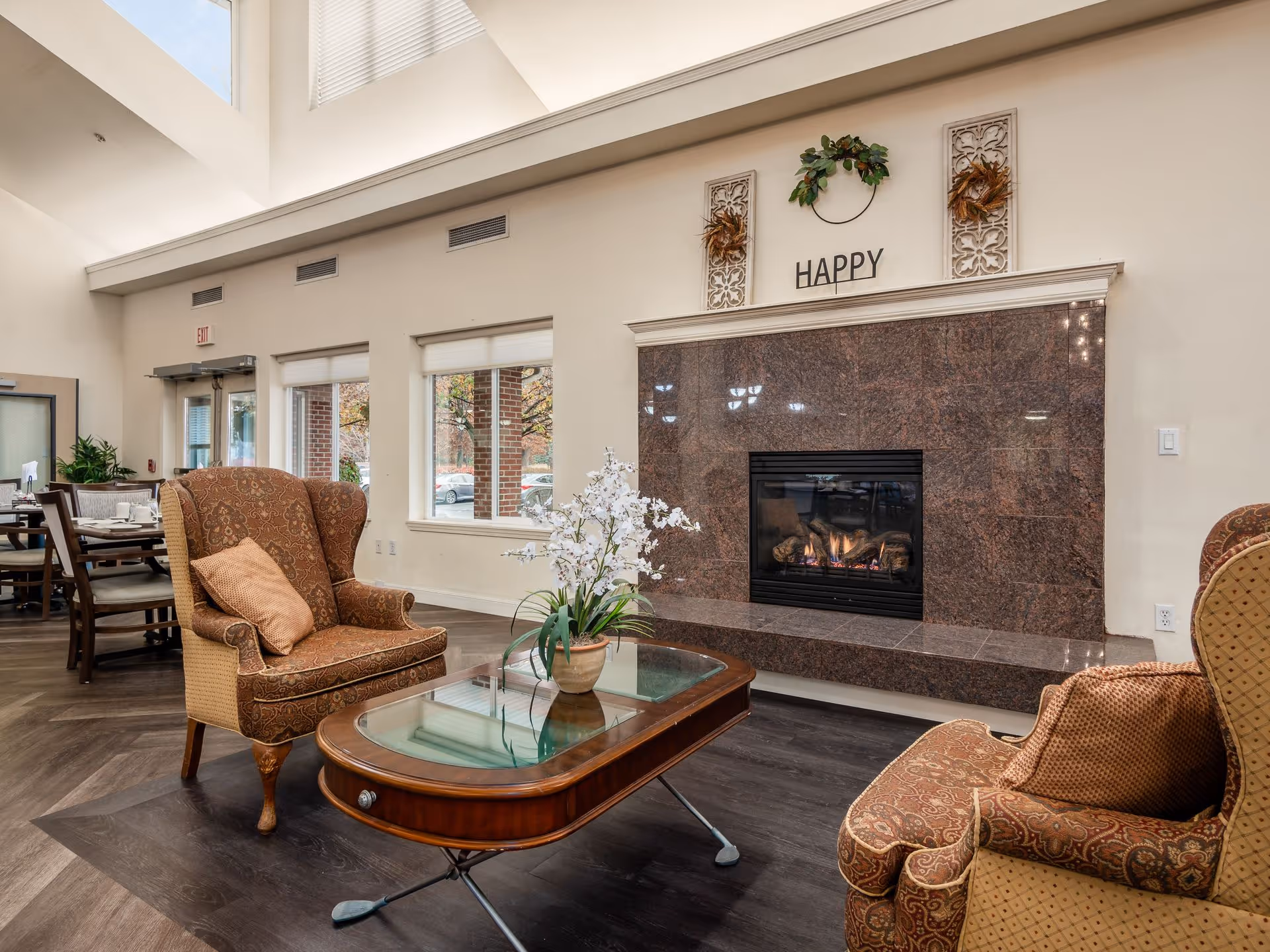 A cozy living room area in a senior living facility featuring two patterned armchairs with cushions, a wooden coffee table with a glass top and a potted white orchid, and a large fireplace with a polished stone surround. Above the fireplace are decorative wall hangings and the word 'HAPPY'. Large windows and a glass door allow natural light to fill the space.