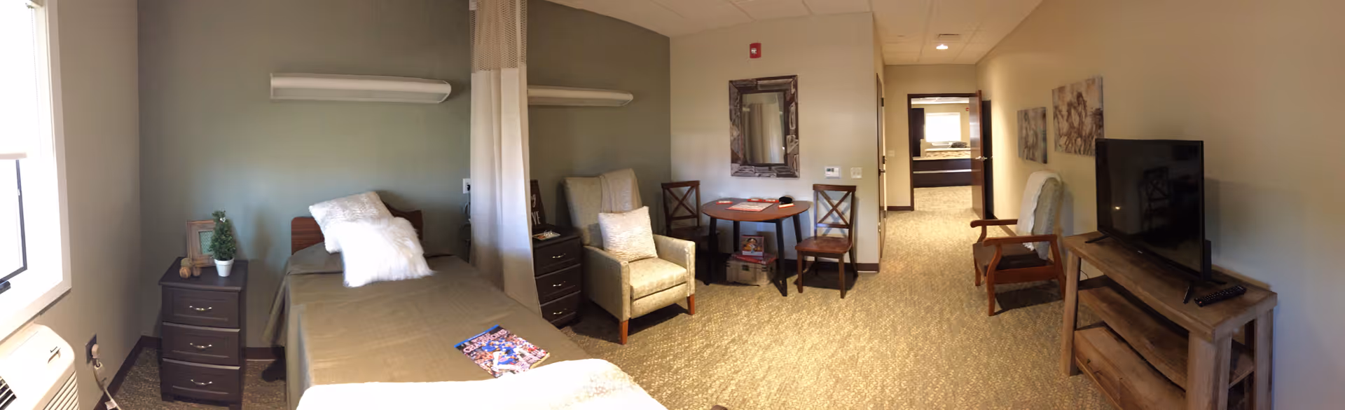 A cozy senior living bedroom with a single bed covered in a brown blanket and a white pillow. Next to the bed is a dark wooden nightstand with a small potted plant and a teddy bear. There is a comfortable armchair and a small round table with two chairs. The room has beige walls and carpeted floor. A flat-screen TV is placed on a wooden TV stand opposite the bed. The room is well-lit with natural light coming from a window on the left.