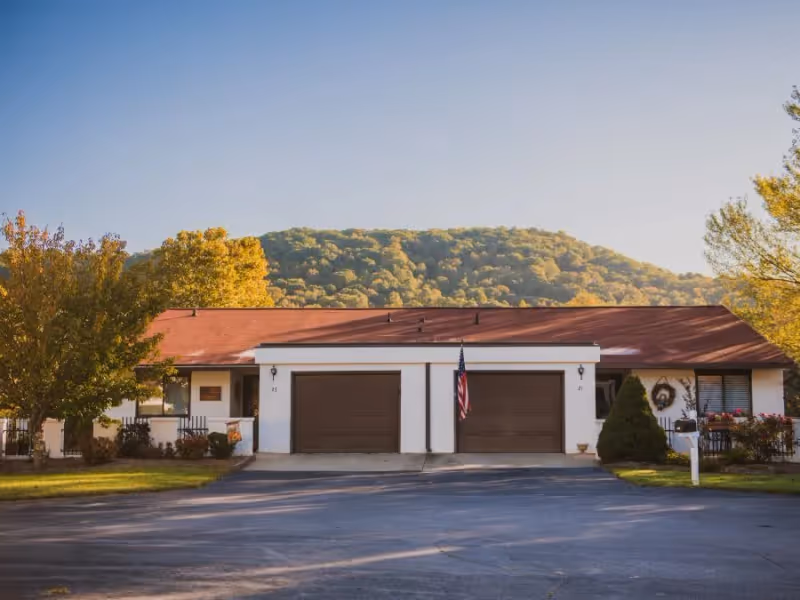 Single-story residential building with two brown garage doors, an American flag hanging between them, surrounded by trees with autumn foliage and a mountain in the background under a clear sky.