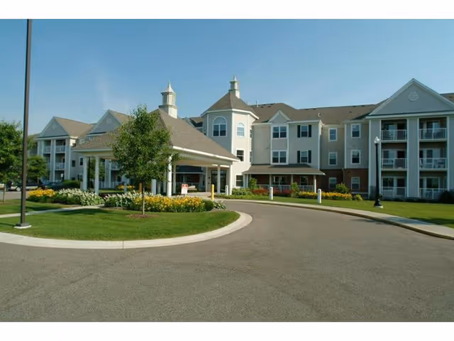 Exterior view of a multi-story senior living facility with a covered entrance, landscaped greenery, and a circular driveway under a clear blue sky.