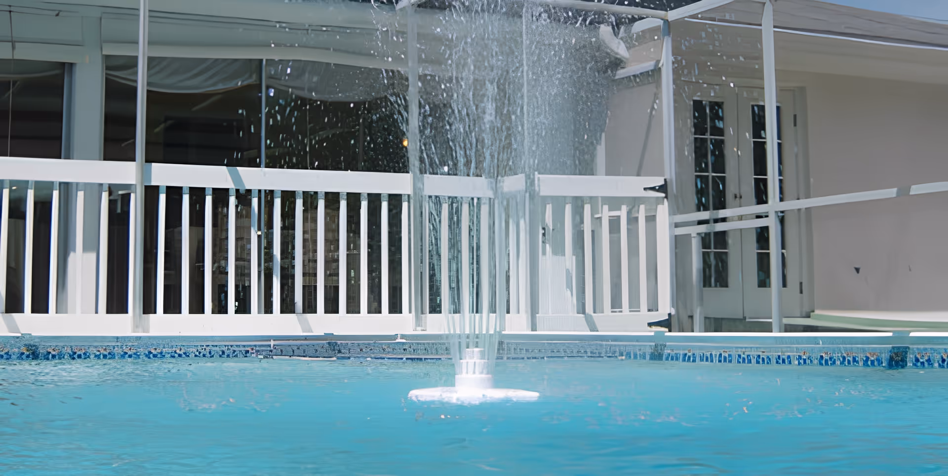 A water fountain spraying water in the middle of a swimming pool with a white railing and building with glass doors in the background.