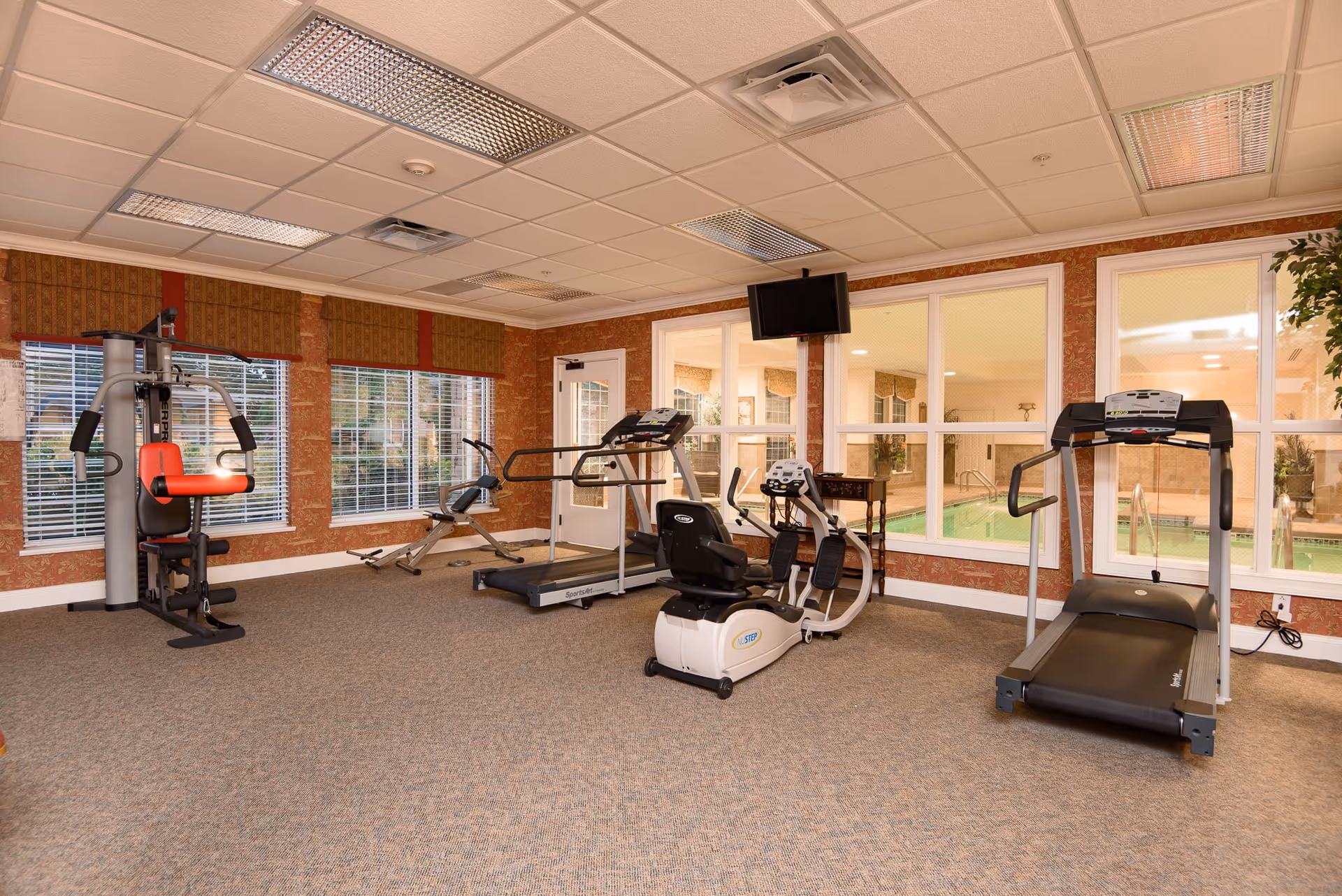 Interior view of a fitness room in a senior living facility with exercise equipment including a multi-gym machine, a rowing machine, a recumbent bike, and two treadmills. The room has large windows with blinds, patterned wallpaper, carpeted floor, and a wall-mounted TV. Through the windows, an indoor swimming pool area is visible.