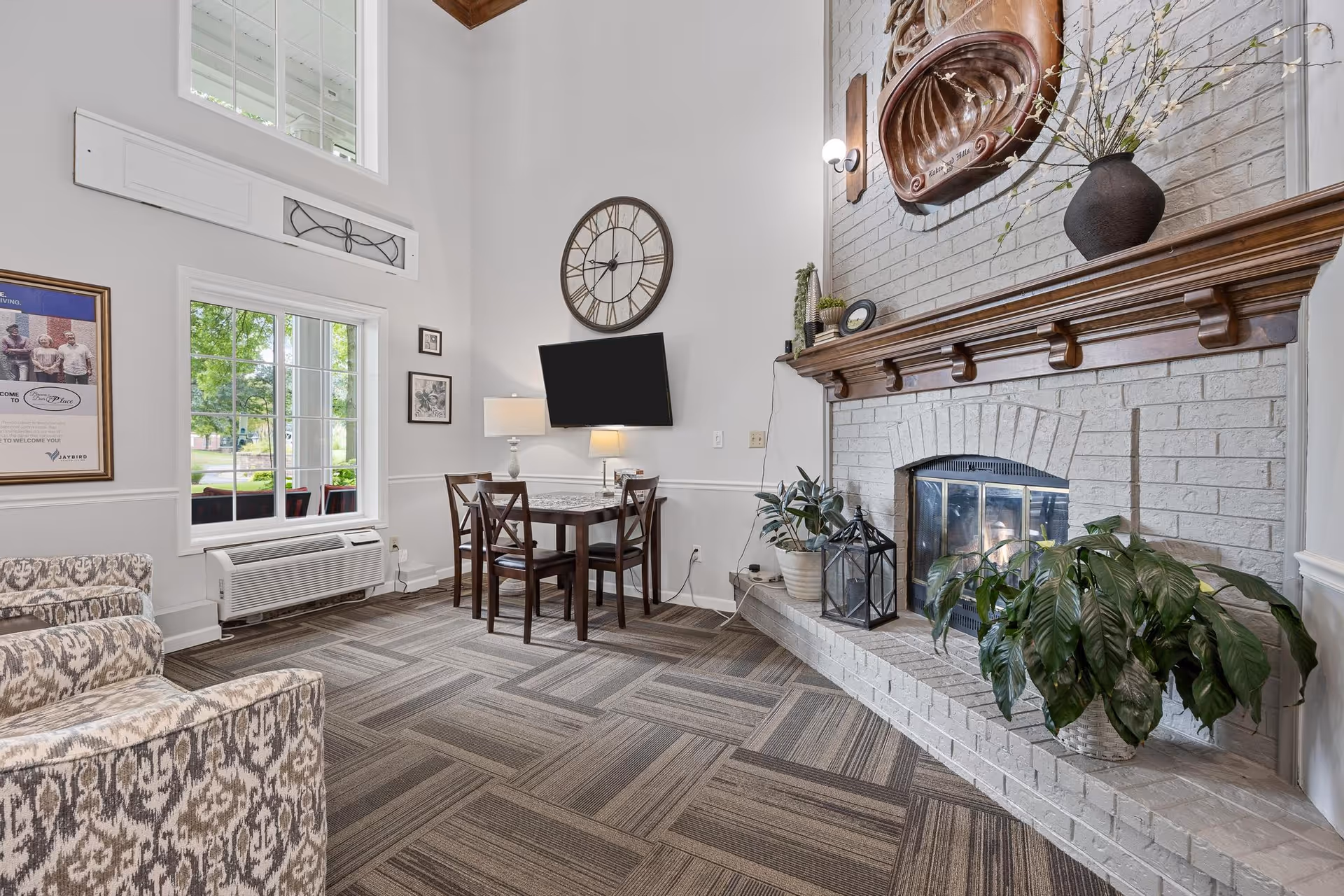 A bright living room area with a large white brick fireplace decorated with plants and a large copper wall hanging. There is a small wooden dining table with four chairs, a wall-mounted TV, a large round clock, and a window showing greenery outside. The room has patterned carpet flooring and light-colored walls with high ceilings.