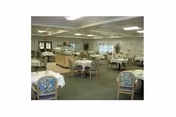 A spacious dining room with multiple round tables covered with white tablecloths and surrounded by chairs with floral upholstery. The room has large windows allowing natural light, and a serving area or counter is visible in the background.