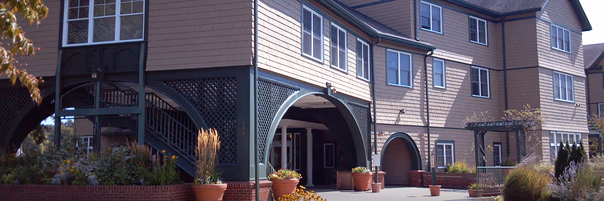 Exterior view of Bertram House of Swampscott showing a multi-story building with beige siding and green trim. The entrance area features large arches, stairs, potted plants, and landscaping with flowers and shrubs.