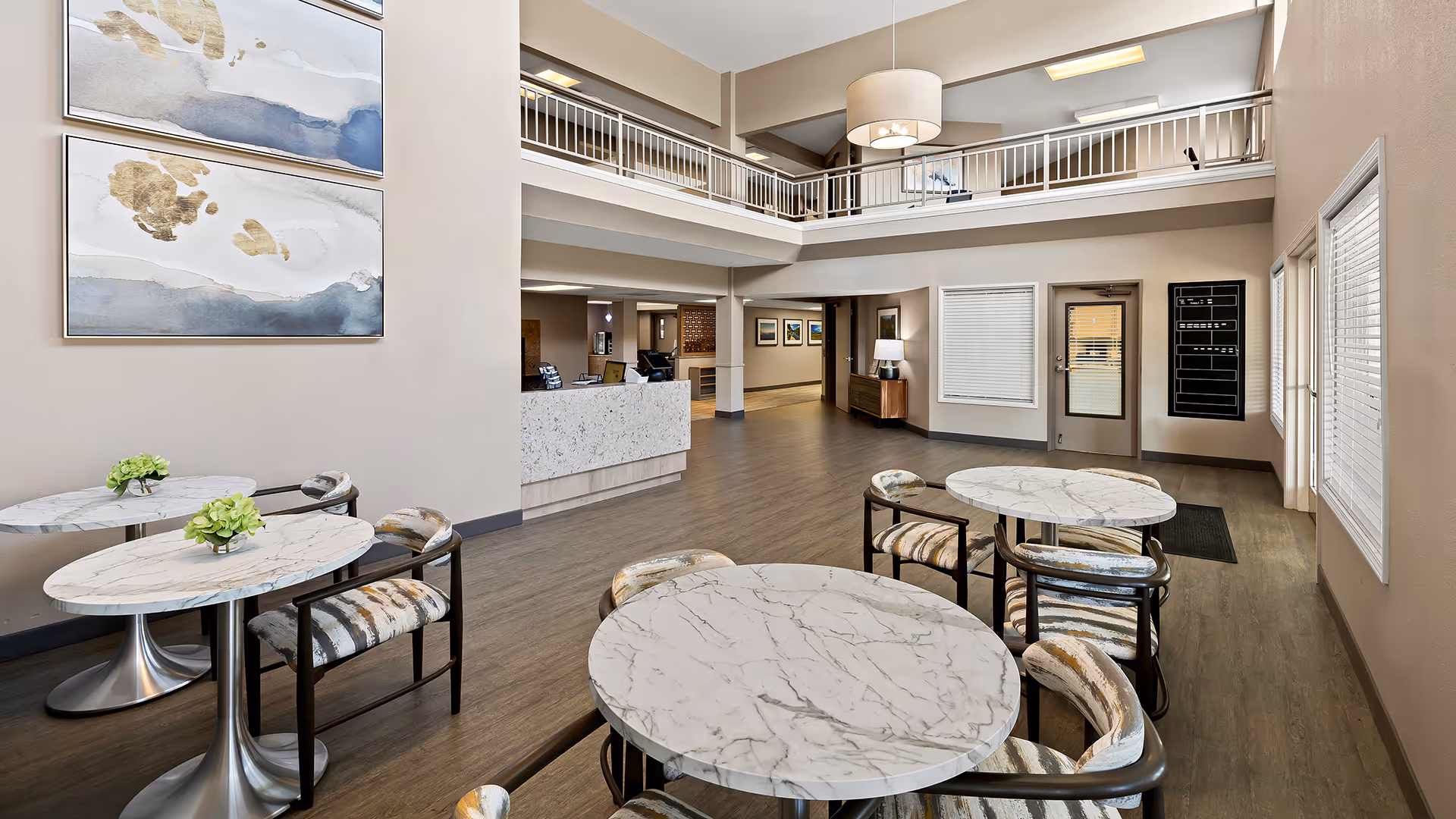 A spacious and well-lit common area with marble-top round tables and cushioned chairs arranged neatly. The room features a high ceiling with a second-floor balcony railing visible. There is a reception desk in the background, modern artwork on the wall, and large windows with blinds allowing natural light to enter.