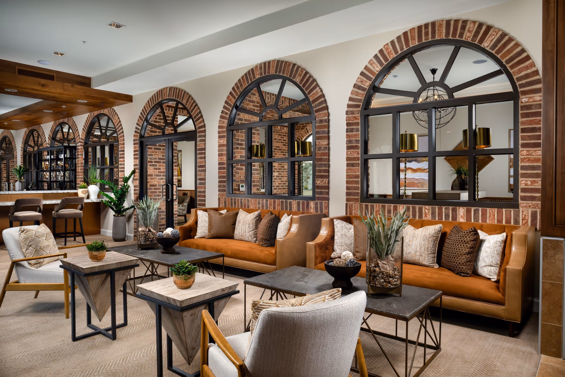 A stylish and cozy living room area featuring two brown leather sofas with multiple decorative pillows, two beige armchairs with patterned cushions, and several modern coffee and side tables adorned with small plants and decorative items. The room has a warm ambiance with brick archways and large black-framed windows and doors, allowing a view into an adjacent room with pendant lighting and artwork.