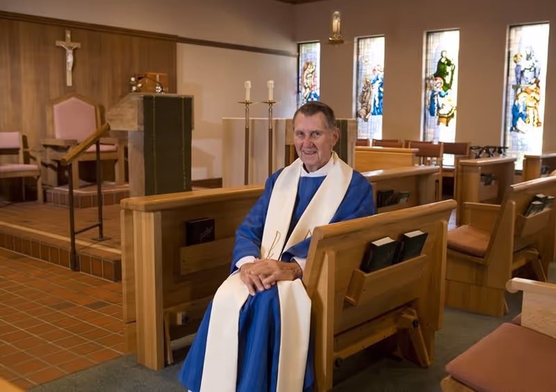 An elderly man dressed in blue religious robes with a white stole sits on a wooden pew inside a chapel. The chapel features stained glass windows, wooden pews with hymnals, a crucifix on the wall, and a lectern with candles.