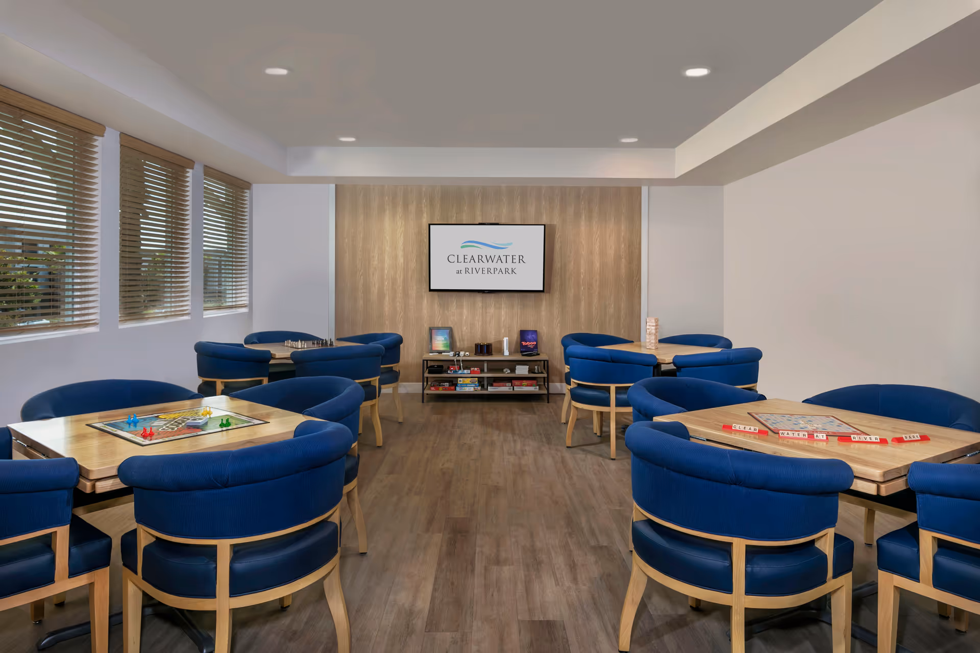 A bright game room with four wooden tables, each surrounded by blue cushioned chairs. Board games such as Scrabble, Sorry, chess, and Jenga are set up on the tables. The room has large windows with wooden blinds on the left side and a wooden accent wall at the far end with a mounted screen displaying the Clearwater at Riverpark logo. Below the screen is a shelf holding additional board games and game accessories.