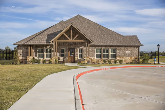 Exterior view of a single-story brick building with a gabled roof and a covered entrance. The building is surrounded by a grassy area and a curved concrete driveway with a red-painted curb. There is a lamppost and a fire hydrant near the driveway.