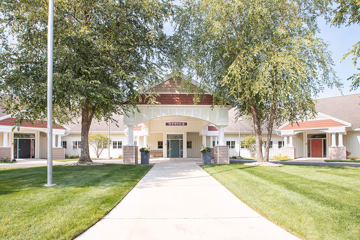 Front entrance of a senior living facility with a walkway leading to an office under a covered portico, flanked by trees and lawns.