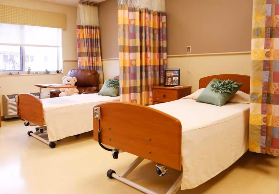 A bright shared senior bedroom with two hospital-style beds, colorful privacy curtains, a recliner, and a bedside table.