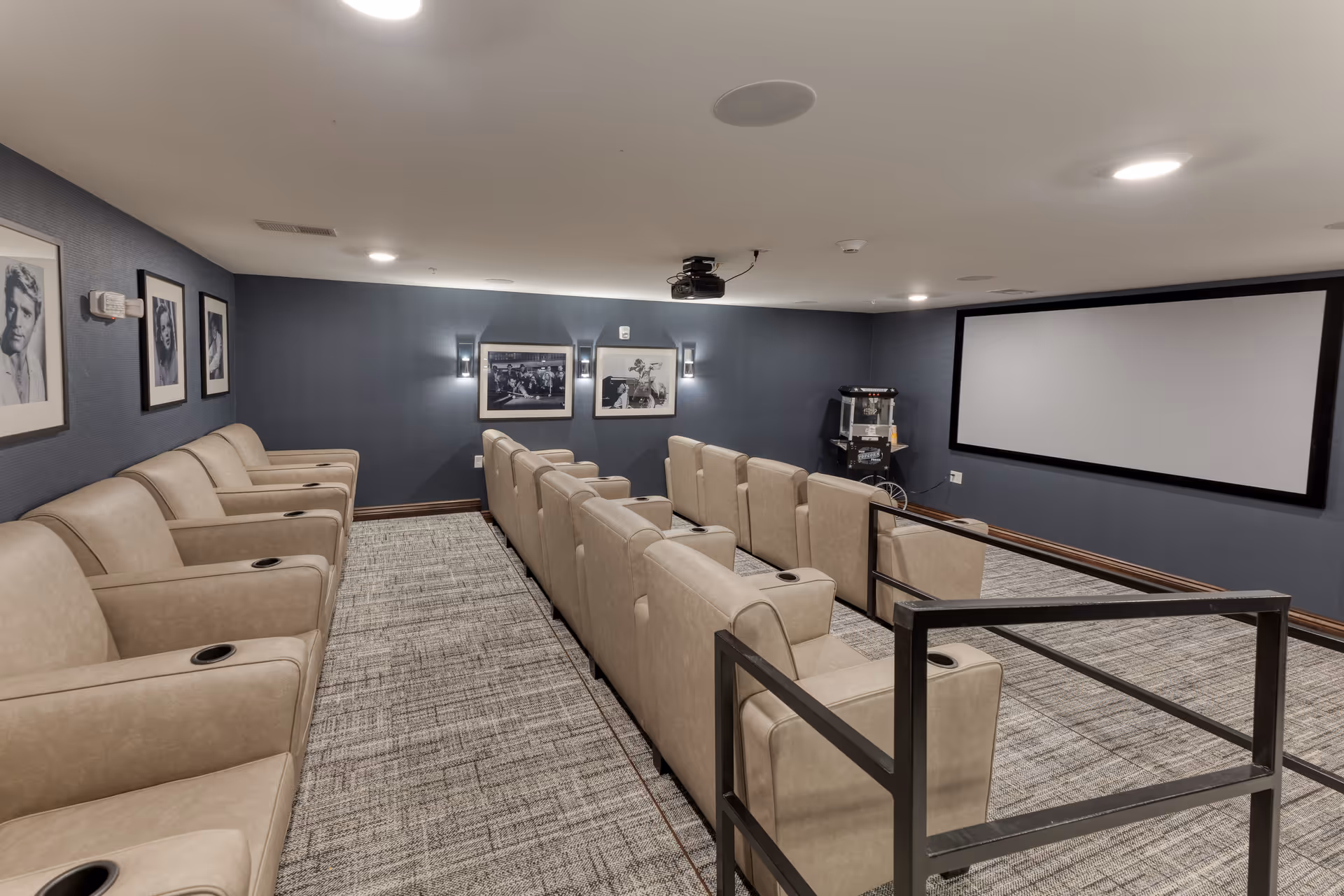 Small private movie theater with two rows of beige recliner seats facing a large projection screen and framed photos on dark blue walls.