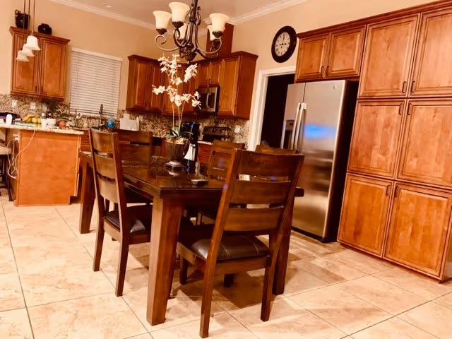 A spacious kitchen and dining area featuring a wooden dining table with six chairs, a chandelier overhead, wooden cabinets, a stainless steel refrigerator, a microwave, and a tiled floor. There is a decorative plant on the dining table and a clock on the wall above the refrigerator.