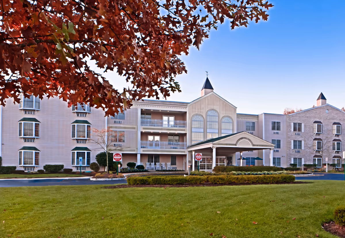 Exterior view of Brandywine Princeton by Monarch senior living facility with a well-maintained lawn and landscaping in the foreground. The building has multiple floors with large windows and a covered entrance. Red autumn leaves from a tree frame the top left corner of the image under a clear blue sky.