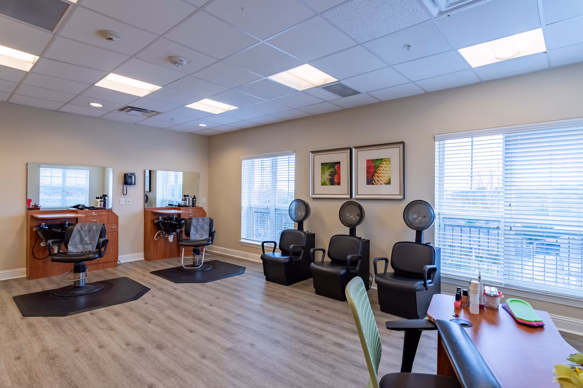 Interior of a salon room with two hair washing stations with chairs and sinks on the left, three black hair drying chairs with hooded dryers along the back wall, two large windows with blinds, framed pictures on the wall, and a manicure table with nail polish and supplies on the right.