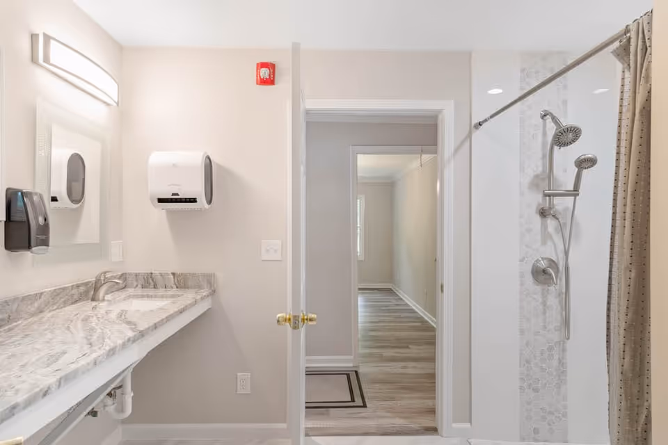 Bright bathroom with a marble vanity and sink, wall-mounted soap and paper towel dispensers, and a tiled walk-in shower seen through an open door.