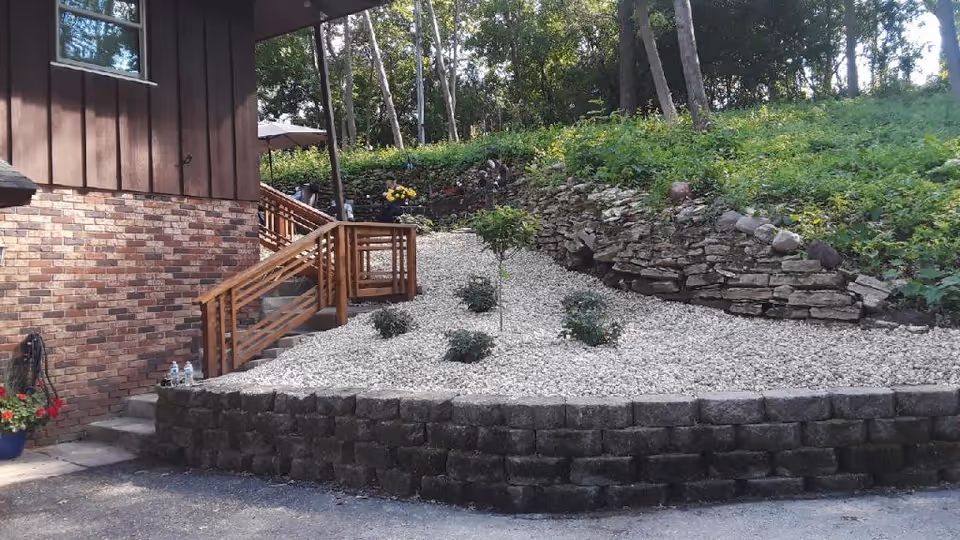Outdoor area beside a building with a brick and wood exterior. There is a small landscaped garden with white gravel, a few small bushes, and a young tree. Stone retaining walls and a wooden staircase with railings lead up to a higher level with more greenery and trees in the background.