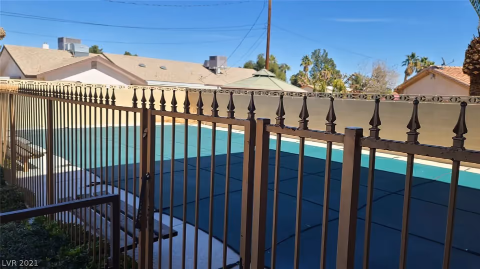 View through a metal fence of a covered outdoor swimming pool with houses and a clear blue sky in the background.
