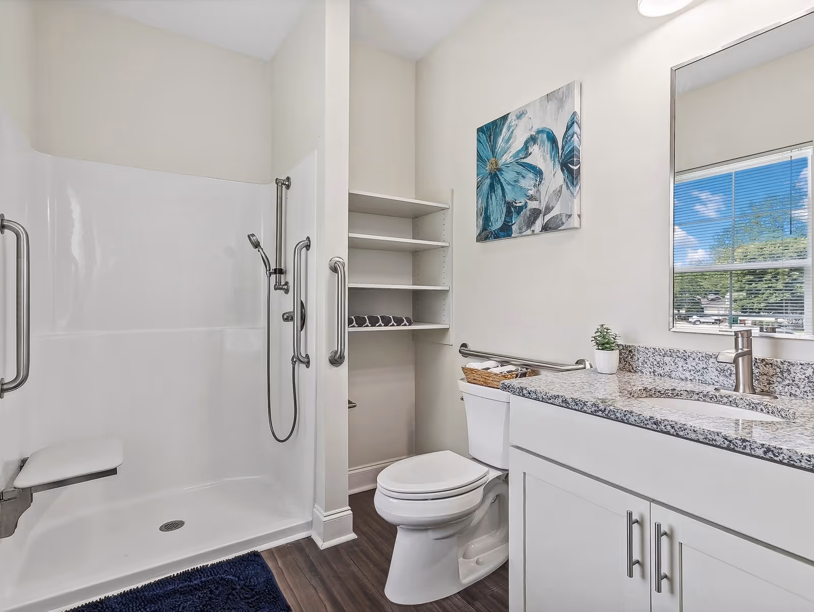 A clean and modern bathroom featuring a walk-in shower with grab bars and a fold-down seat, a toilet with a grab bar beside it, white cabinetry with a granite countertop and sink, a small potted plant, a basket with towels, open shelving with folded towels, a large mirror, and a window showing a blue sky with clouds outside. A blue floral painting hangs on the wall above the toilet.