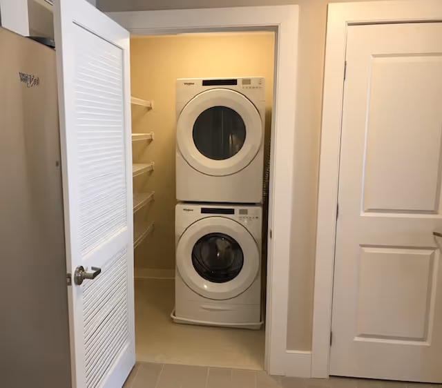 Laundry area with a stacked white Whirlpool washer and dryer inside a small closet with open louvered door. Shelving is visible on the left side inside the closet.