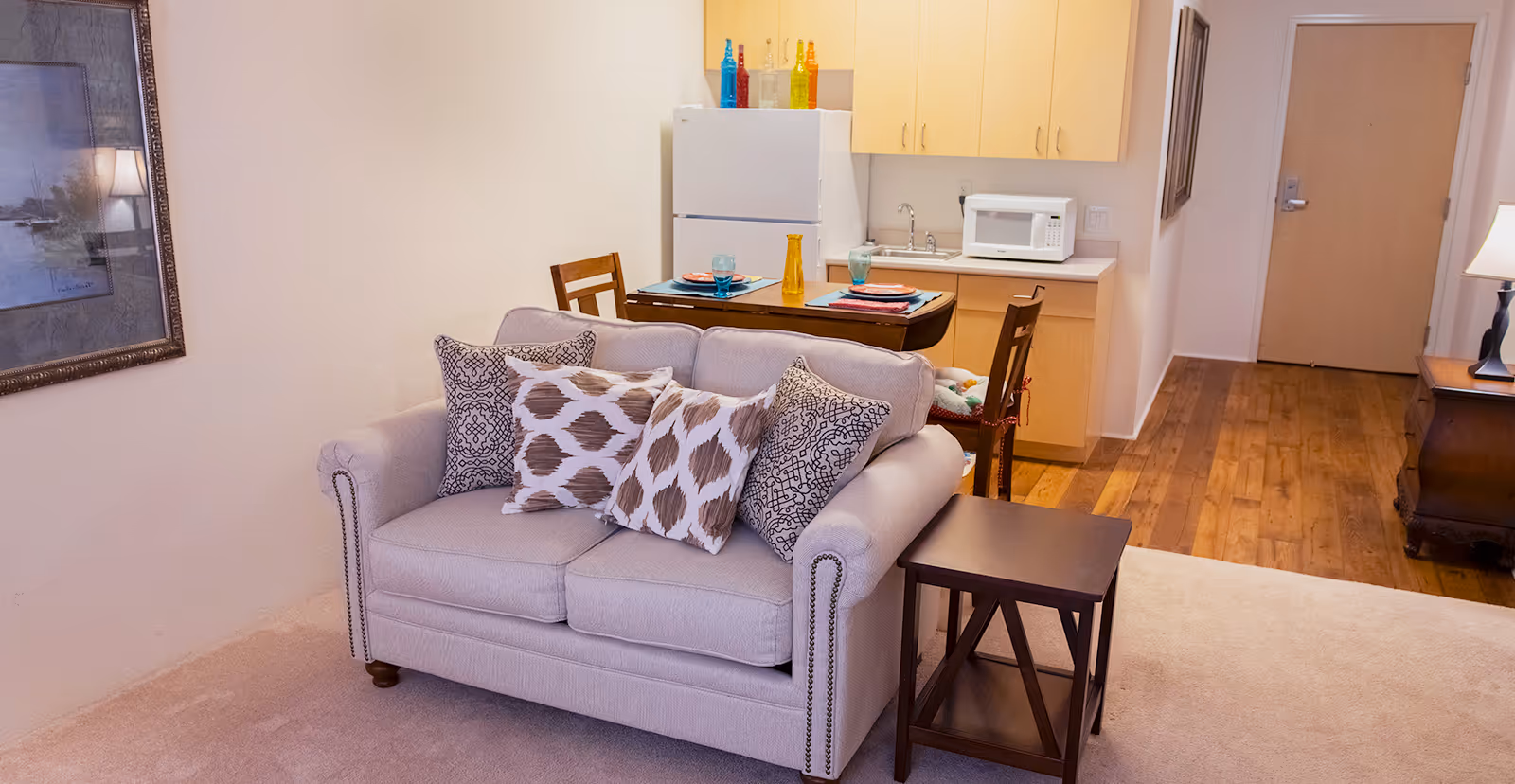 Interior view of a senior living facility showing a small living area with a beige loveseat adorned with patterned throw pillows, a wooden side table, and a dining area with a table set for two. The background includes a compact kitchen with a refrigerator, microwave, and wooden cabinets. The floor transitions from carpet in the living area to wood in the hallway leading to a closed door.