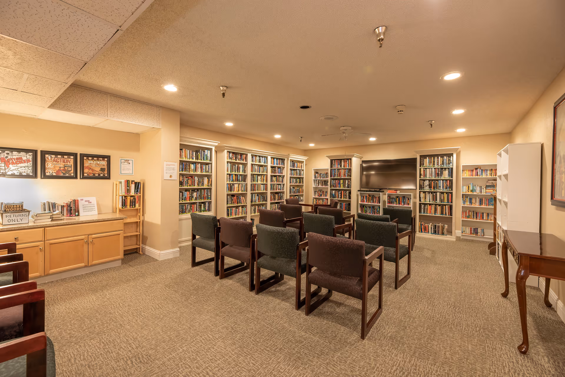 A cozy library room with multiple bookshelves filled with books along the walls. Several chairs are arranged in rows facing a large flat-screen TV mounted on the wall. The room has warm lighting and a carpeted floor, with framed pictures and a wooden cabinet on the left side.