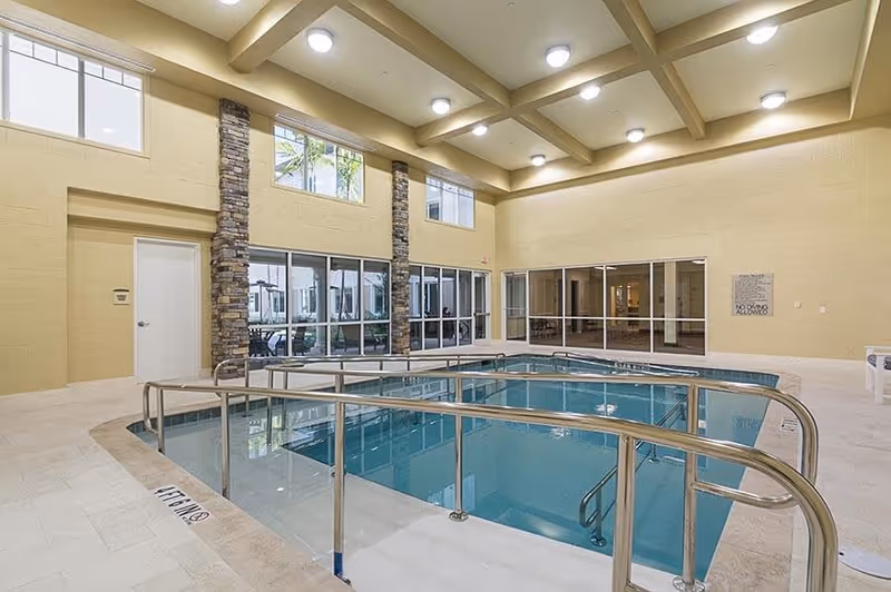 Indoor swimming pool area with handrails and steps leading into the water, surrounded by beige tiled flooring and walls with stone pillars. Large windows and glass doors provide natural light and views of an outdoor area. The ceiling has exposed beams with multiple round lights.