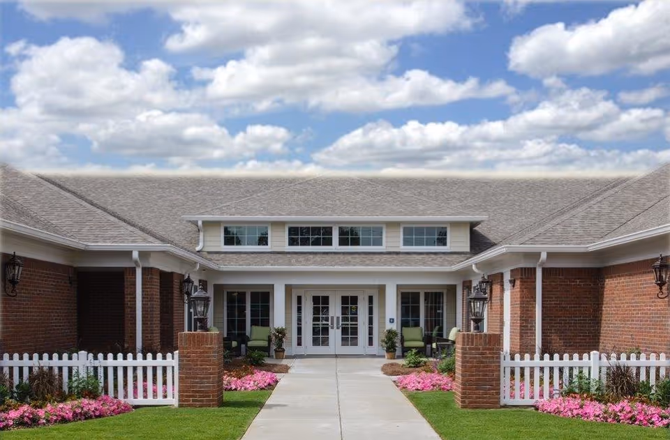 Front entrance of a brick assisted living facility with a central walkway, white picket fences, and flowerbeds.
