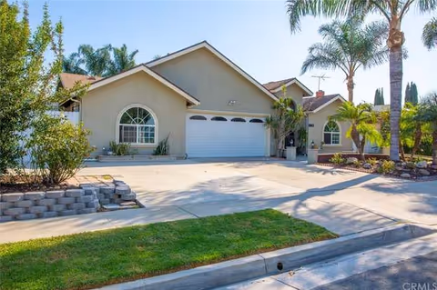 Single-story residential building with a beige exterior, a two-car garage, and arched windows. The driveway is concrete and there are palm trees and other plants around the property under a clear blue sky.