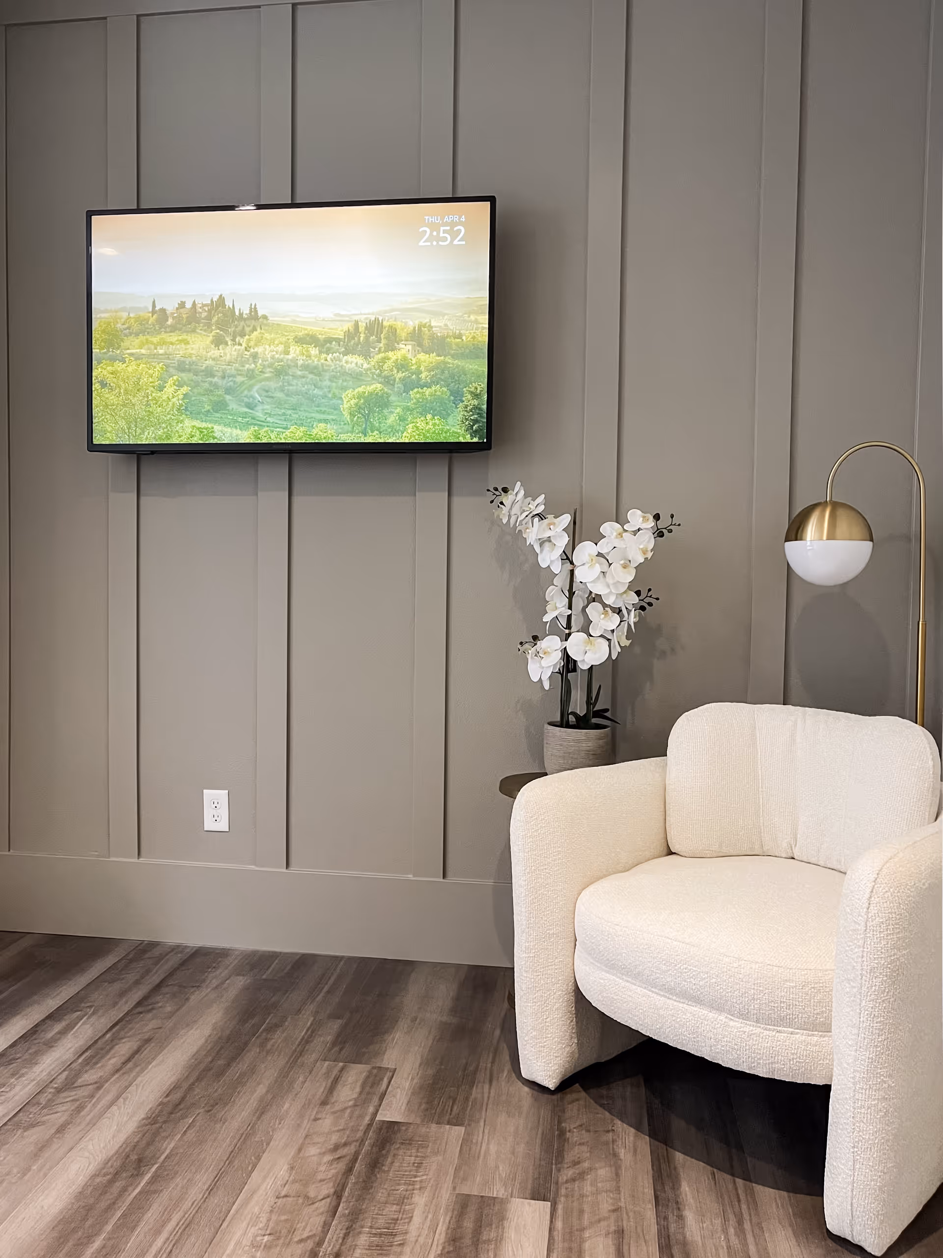 A cozy corner of a living room with a cream-colored armchair, a small side table with a white orchid plant, a modern floor lamp with a gold and white design, and a wall-mounted flat-screen TV displaying a scenic landscape with trees and hills.