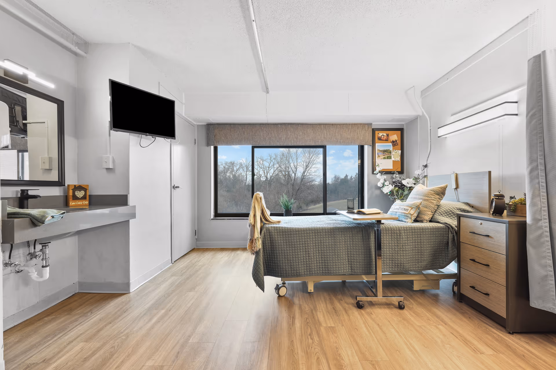 A bright and clean senior living bedroom with a hospital-style bed covered in a gray quilt, a wooden nightstand with drawers, a mounted flat-screen TV, a large window with a view of trees outside, a sink with a mirror on the left wall, and light wood flooring.