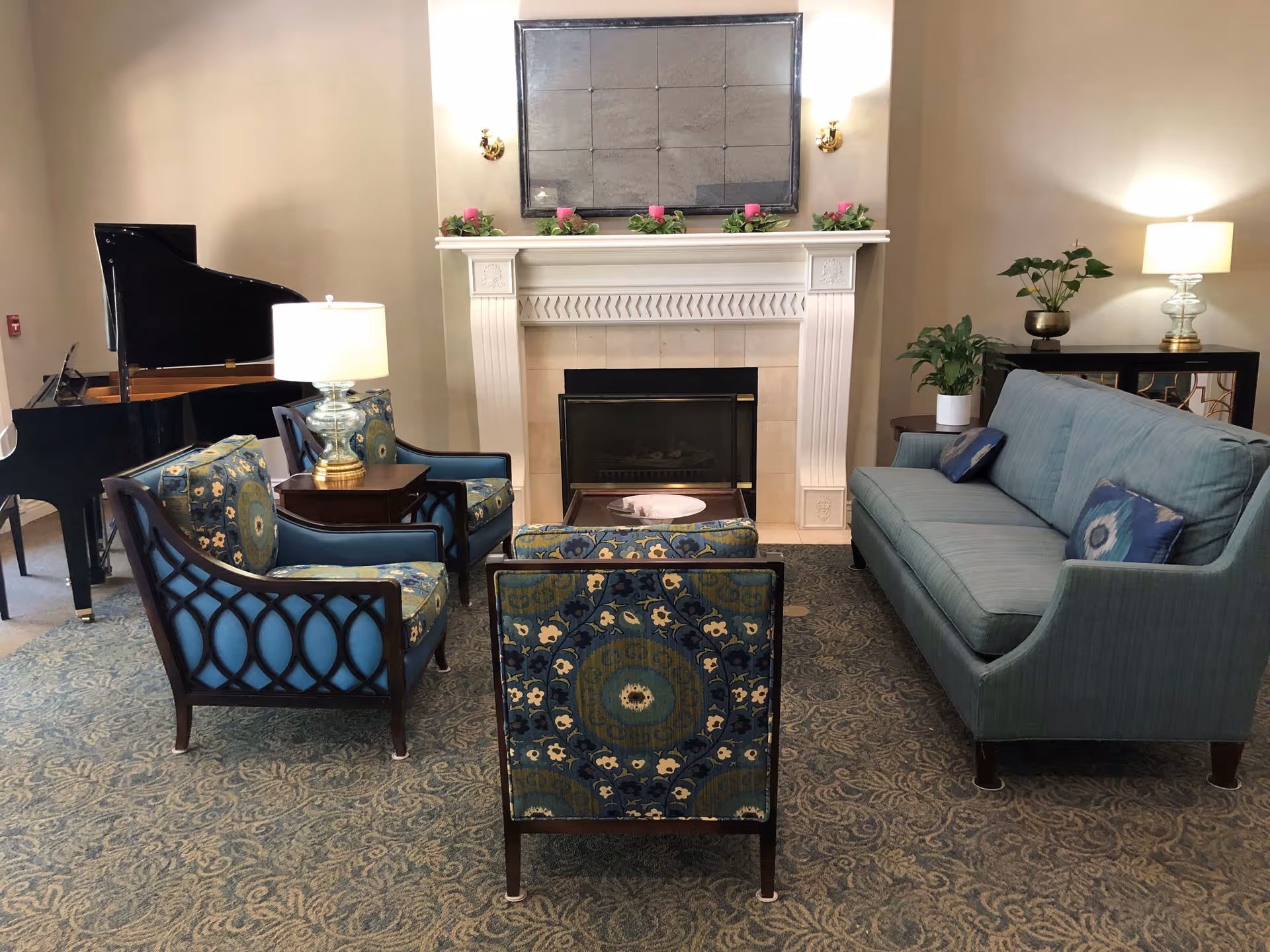 A cozy living room with a blue sofa, three patterned armchairs, a wooden coffee table, and a fireplace with a large mirror above it. There are two table lamps on side tables, plants on a black console table, and a grand piano in the corner.