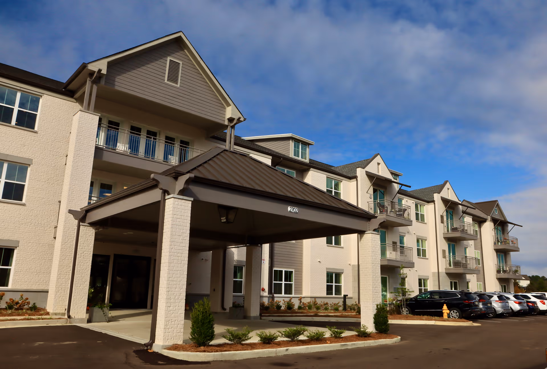 Exterior view of a multi-story senior living facility building with a covered entrance, balconies, multiple windows, and a parking lot with several cars parked. The sky is partly cloudy.