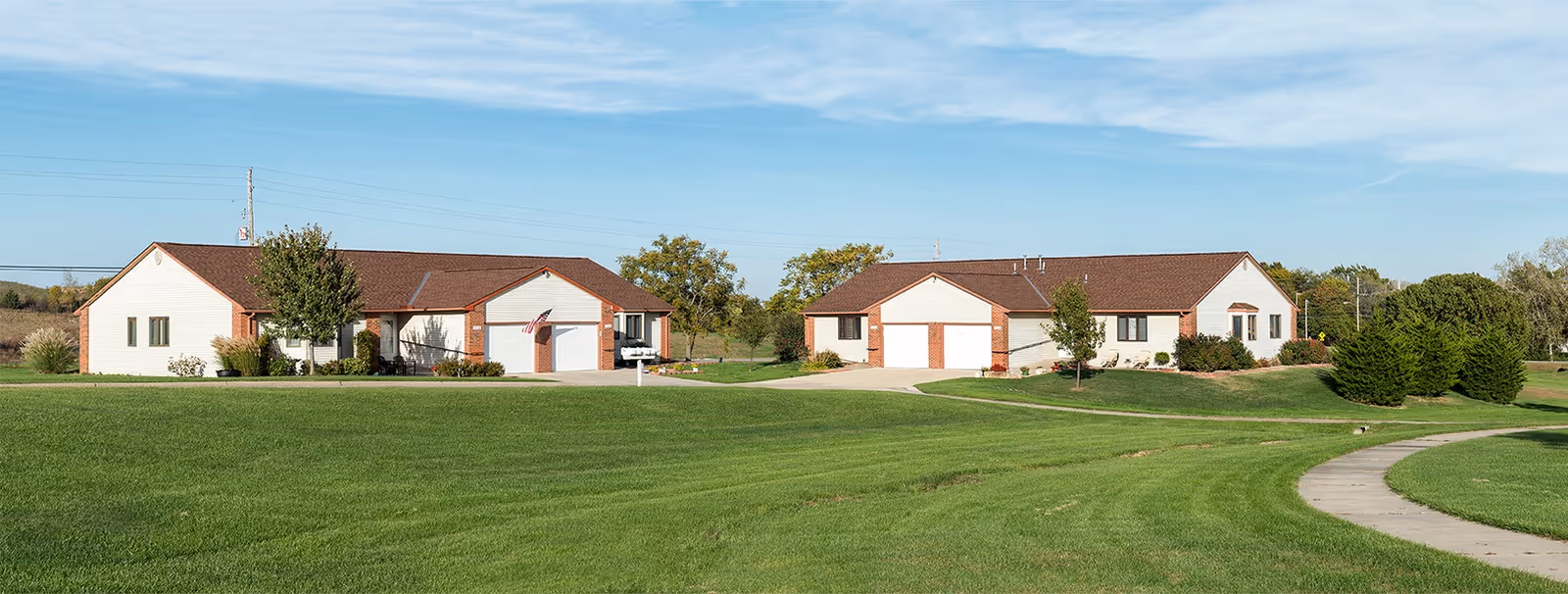 Two single-story buildings with brown roofs and white walls situated on a large green lawn under a blue sky with some clouds. There is a curved concrete pathway on the right side of the image and some trees and shrubs around the buildings.