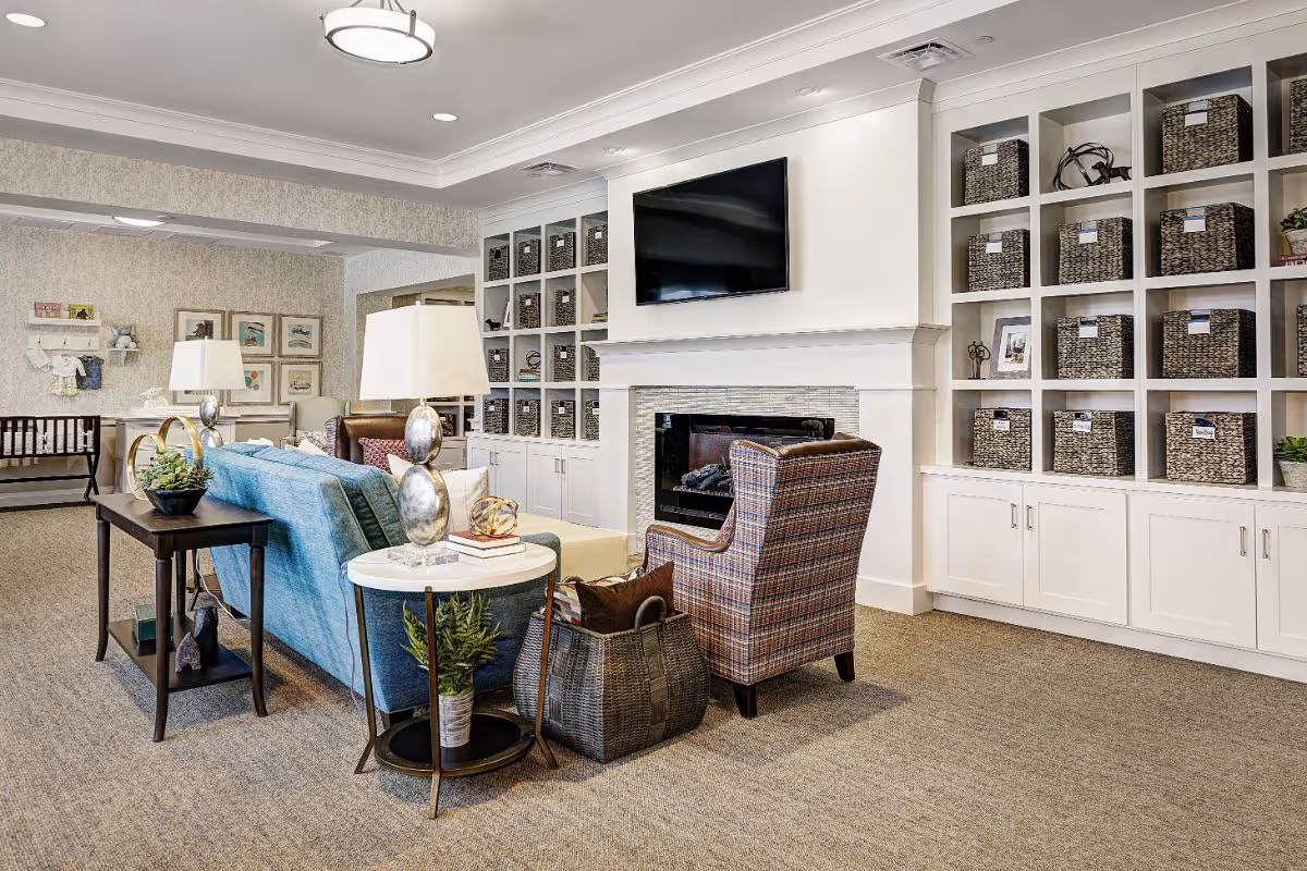 A cozy living room area in a senior living facility featuring a blue sofa, a patterned armchair, a fireplace with a mounted flat-screen TV above it, and built-in white shelves filled with woven baskets. The room is well-lit with ceiling lights and table lamps, and there are decorative plants and books on side tables.