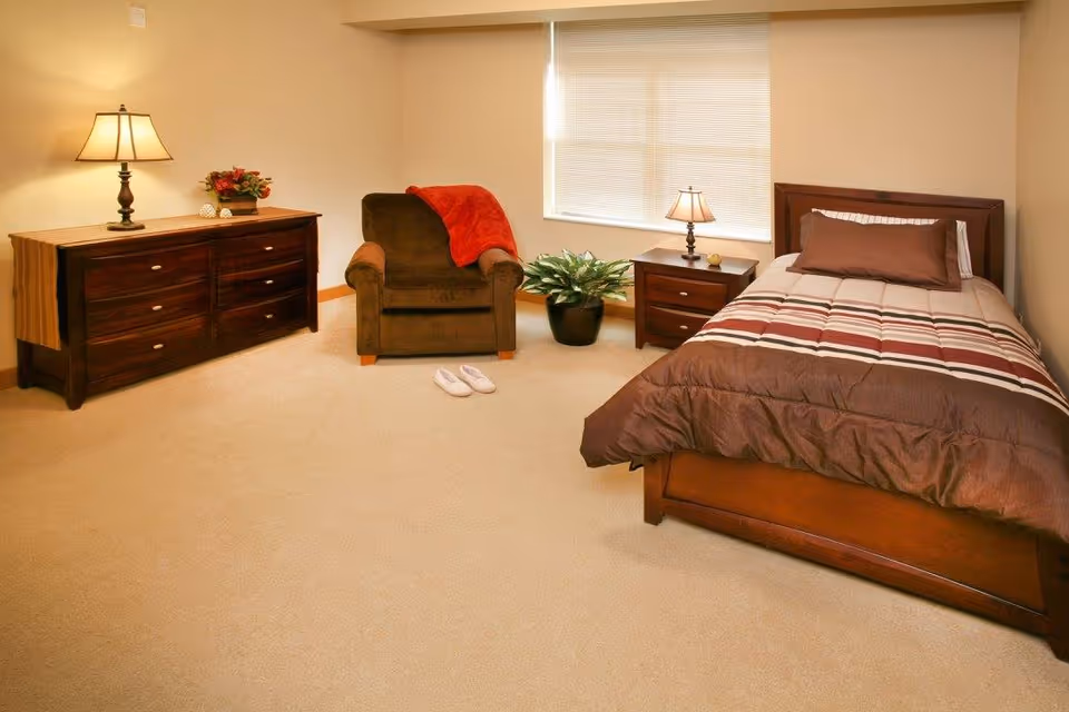 A cozy bedroom with a single bed featuring a striped brown and beige comforter and a brown pillow. Next to the bed is a wooden nightstand with a small lamp and a decorative item. A brown armchair with a red throw blanket is positioned near a window with closed blinds. Across from the chair is a wooden dresser with a lamp and a flower arrangement on top. A pair of white slippers is placed on the beige carpeted floor.
