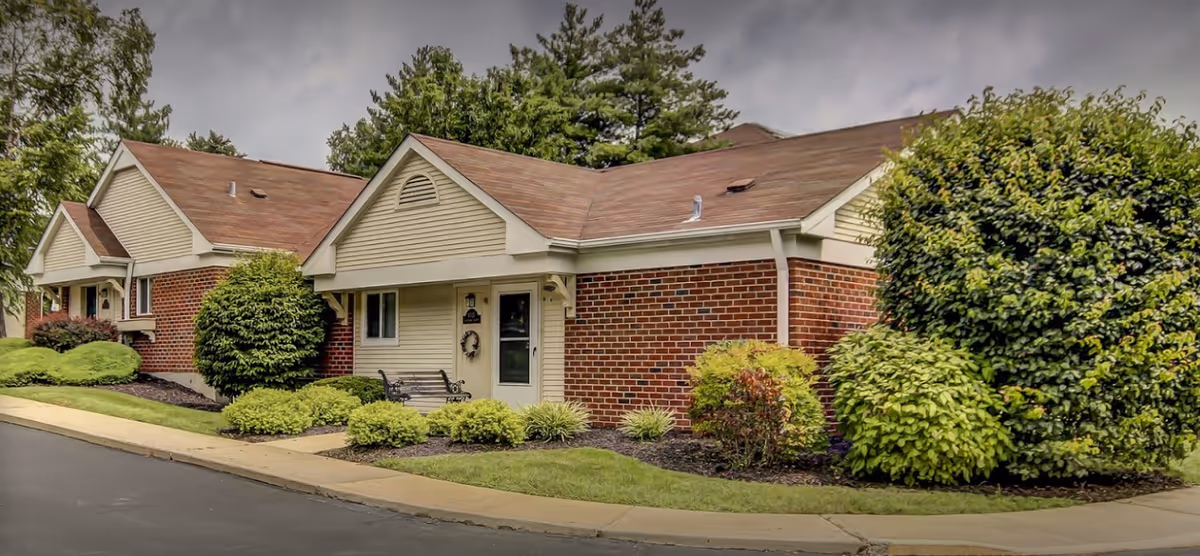 Exterior view of a single-story brick and siding building with a brown shingled roof, surrounded by well-maintained bushes and greenery, with a sidewalk and paved road in front.