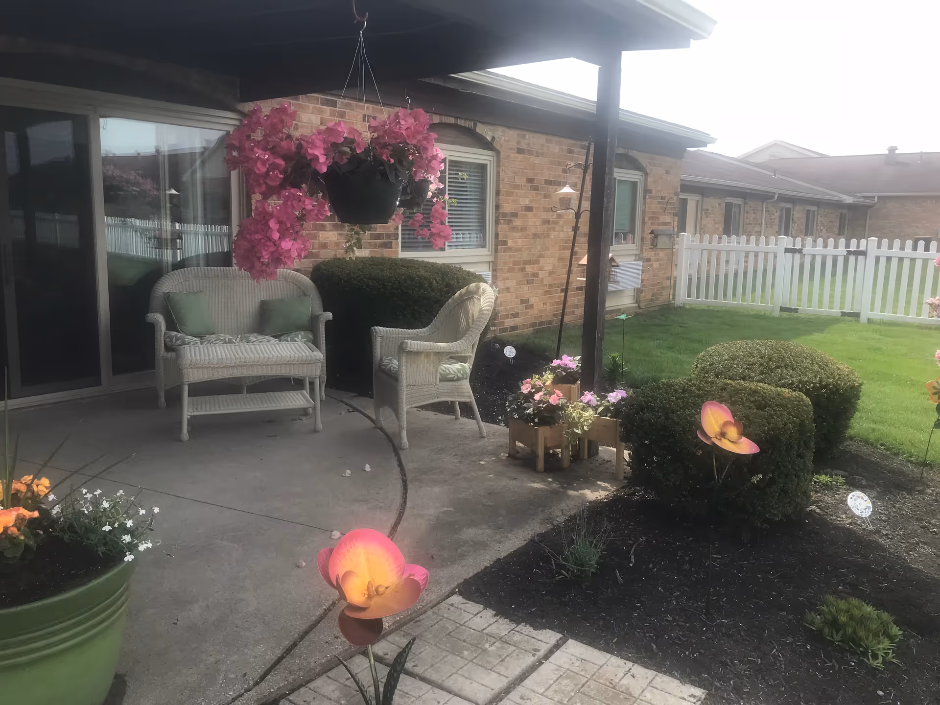 Outdoor patio area with white wicker chairs and a loveseat with green cushions, hanging basket with pink flowers, potted plants, trimmed bushes, and a white picket fence surrounding a grassy yard next to a brick building.