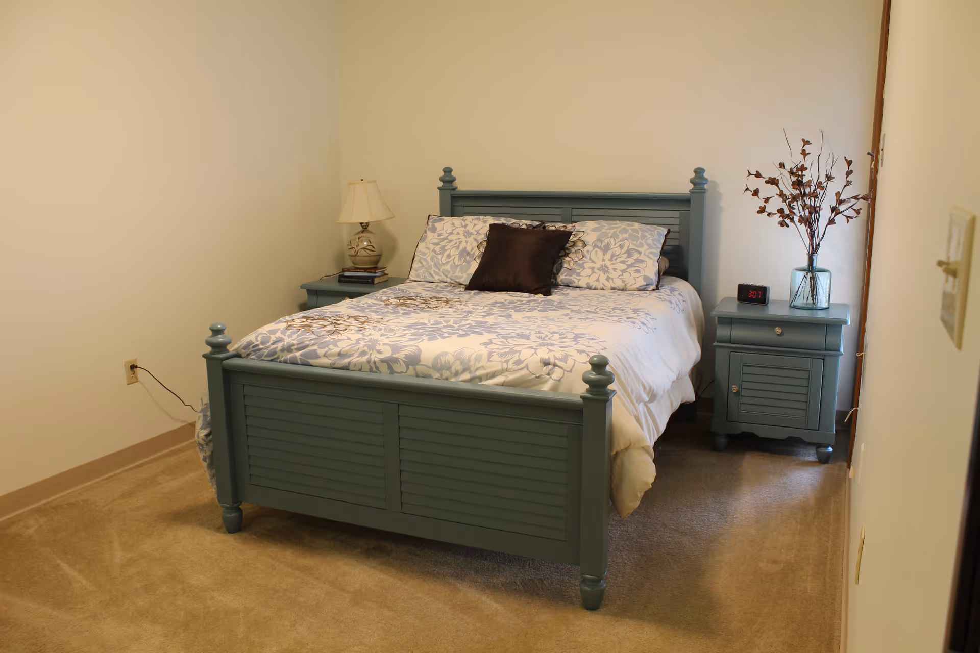 A bedroom with a blue wooden bed frame and matching nightstands on either side. The bed is made with white and blue floral bedding and has three pillows, including one dark brown pillow in the center. One nightstand has a lamp and some books, while the other has a digital clock and a vase with dried branches. The room has beige carpet and plain light-colored walls.