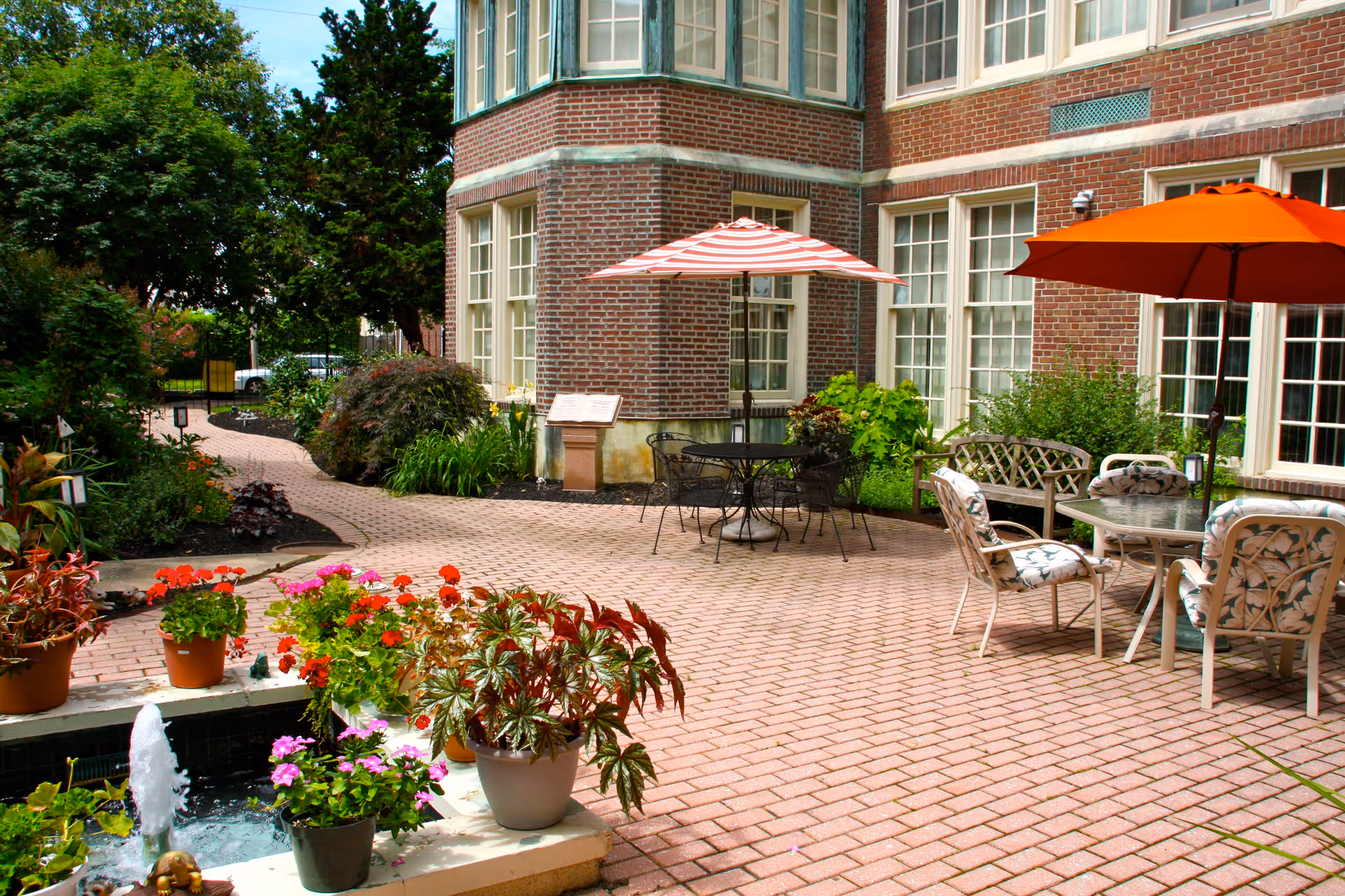 Outdoor patio area at The Lorelton with brick paving, several tables with umbrellas, cushioned chairs, a wooden bench, potted plants, and a small water fountain surrounded by colorful flowers. The patio is adjacent to a brick building with large windows and is bordered by green shrubs and trees.