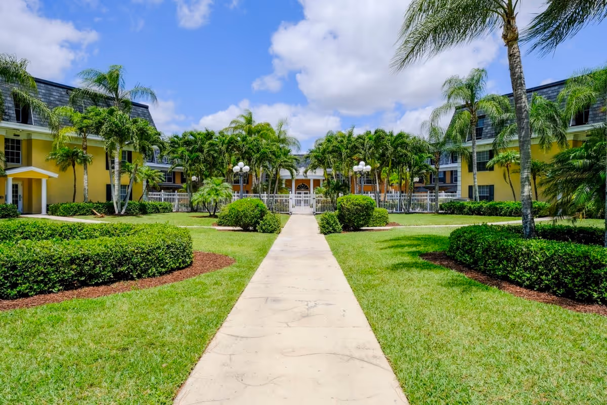 A paved walkway leads through a well-maintained garden with trimmed bushes and palm trees towards a yellow two-story building with a slate roof under a partly cloudy blue sky.
