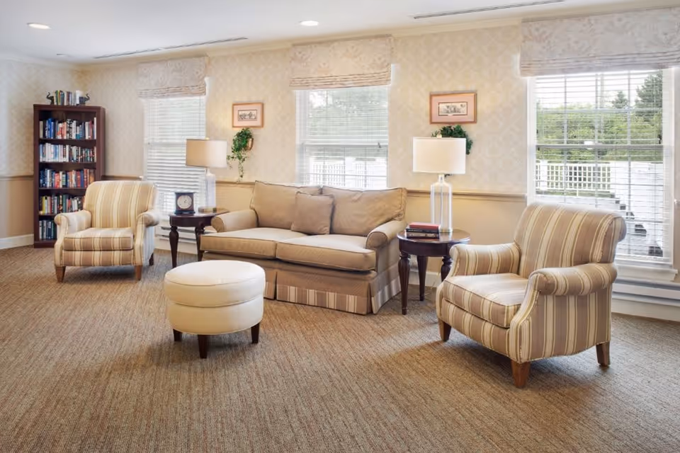 A cozy living room with two striped armchairs, a beige sofa with cushions, a round ottoman, two side tables with lamps and books, a bookshelf filled with books, and three large windows with blinds letting in natural light.