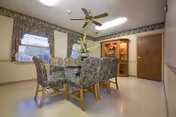 Small dining room with a round table surrounded by floral-upholstered chairs, a china cabinet, ceiling fan, and two windows.