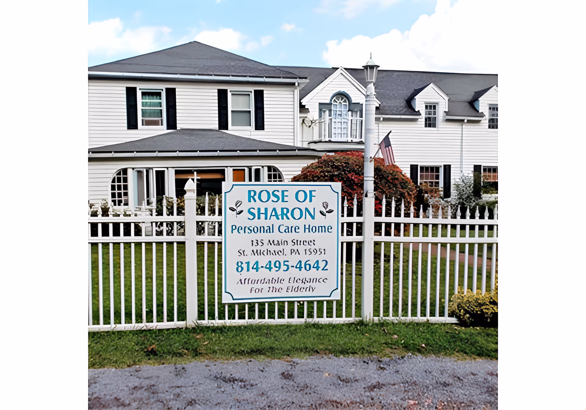 Exterior view of Rose of Sharon Personal Care Home, a white two-story building with black shutters and a gray roof, behind a white picket fence with a sign displaying the facility's name, address, phone number, and slogan.