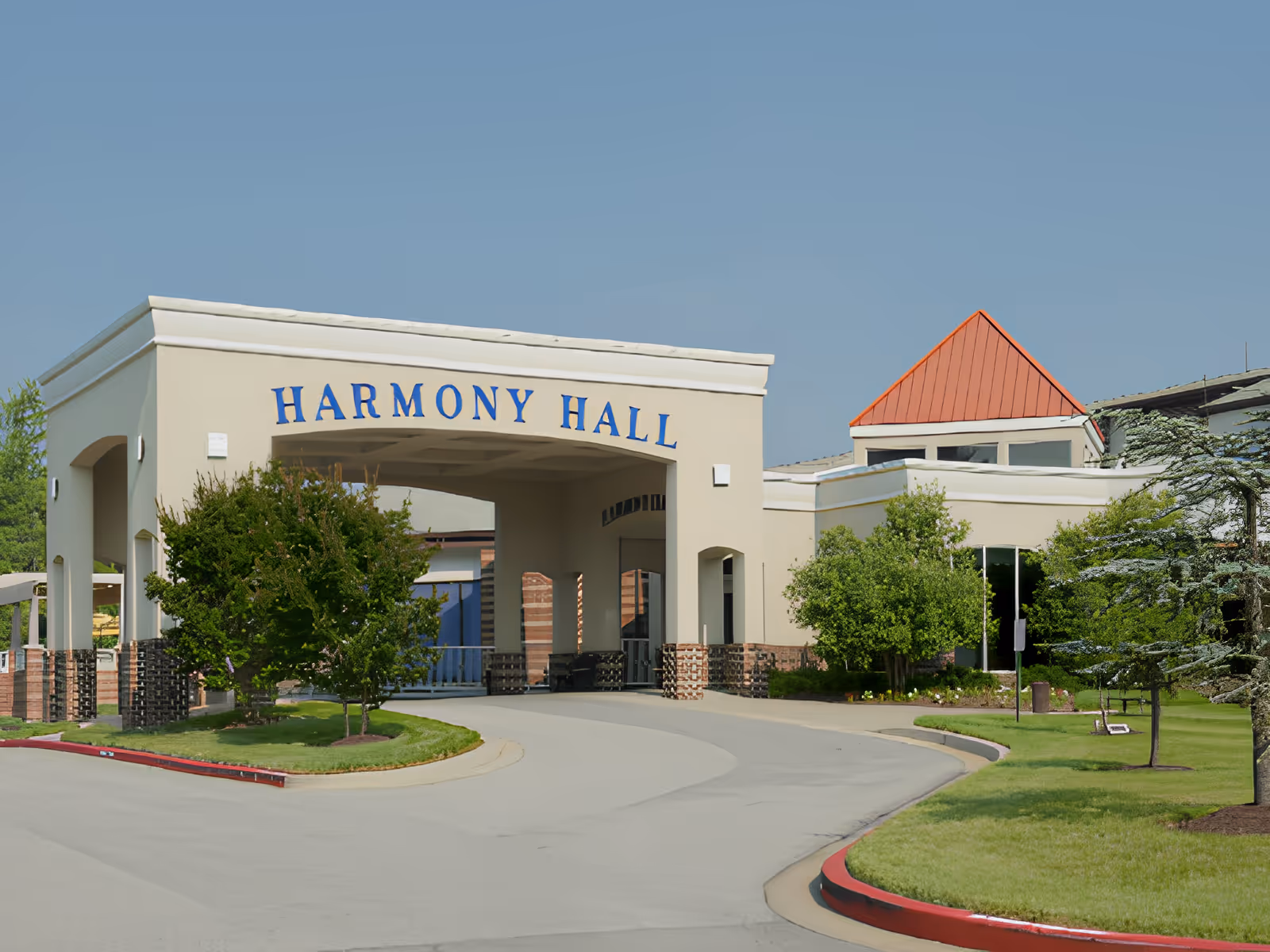 Exterior view of Harmony Hall building with a covered entrance, surrounded by green trees and a well-maintained lawn under a clear blue sky.