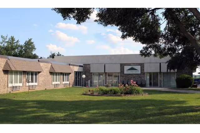 Exterior view of Ambassador Health of Sidney facility showing a single-story building with brick and light-colored walls, several windows, a central entrance with glass doors, and a landscaped lawn with a flower bed in front under a partly cloudy sky.