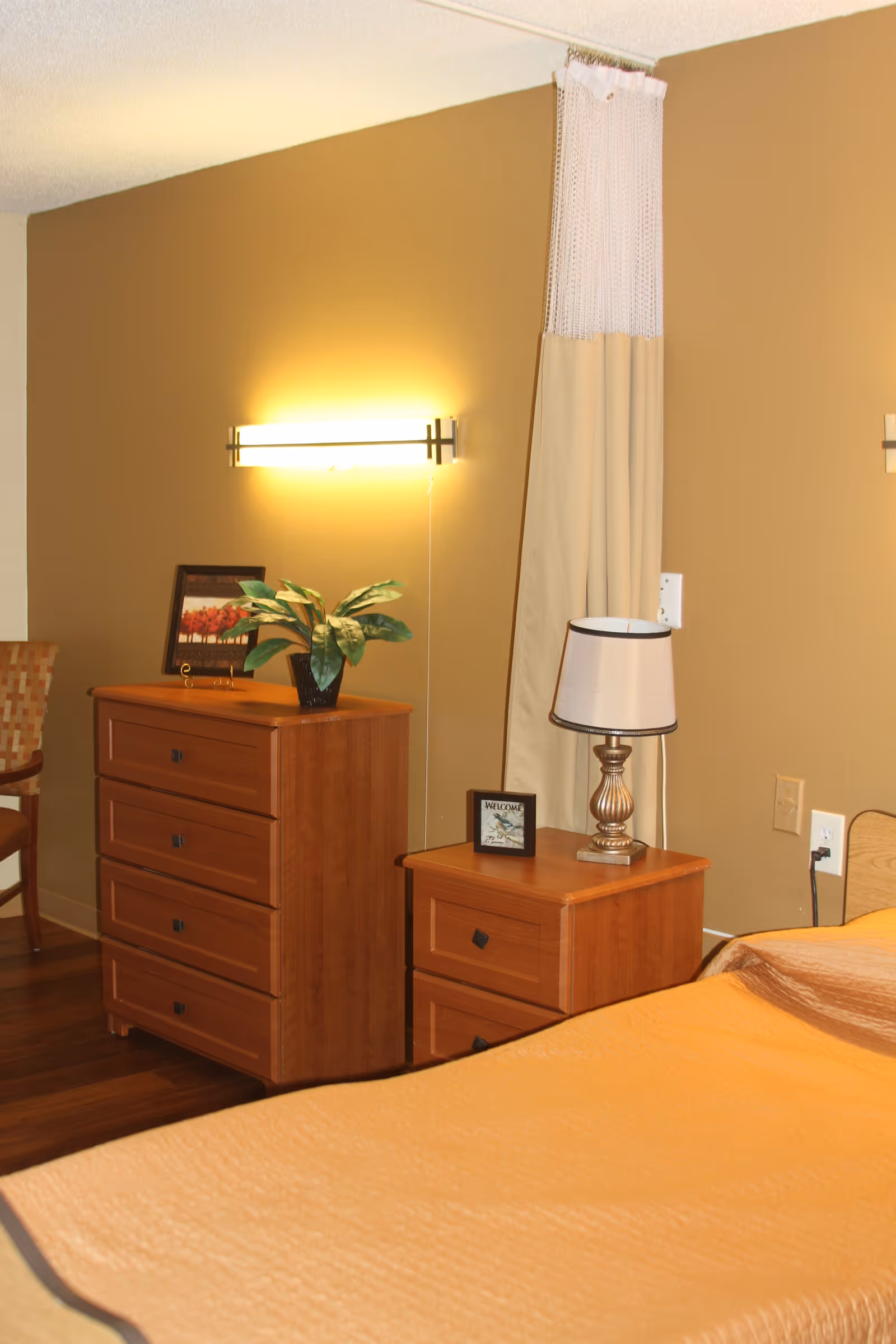Bedroom corner with a wooden dresser and nightstand, lamp, potted plant, and a bed with beige bedding against tan walls.